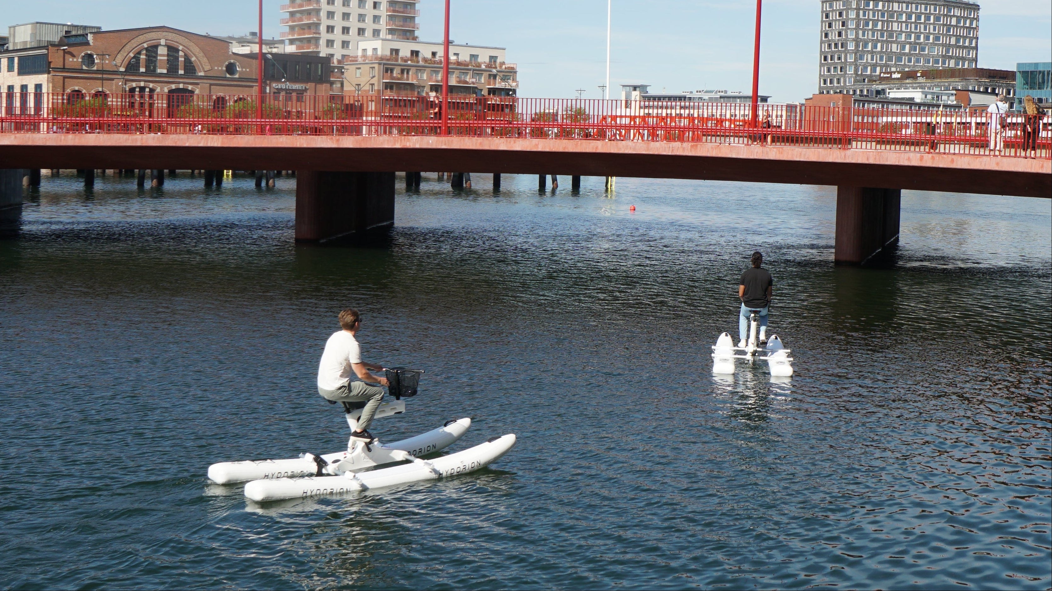 A person stands on a white water bike in the water near a red bridge over a harbor, with city buildings and a blue sky in the background.