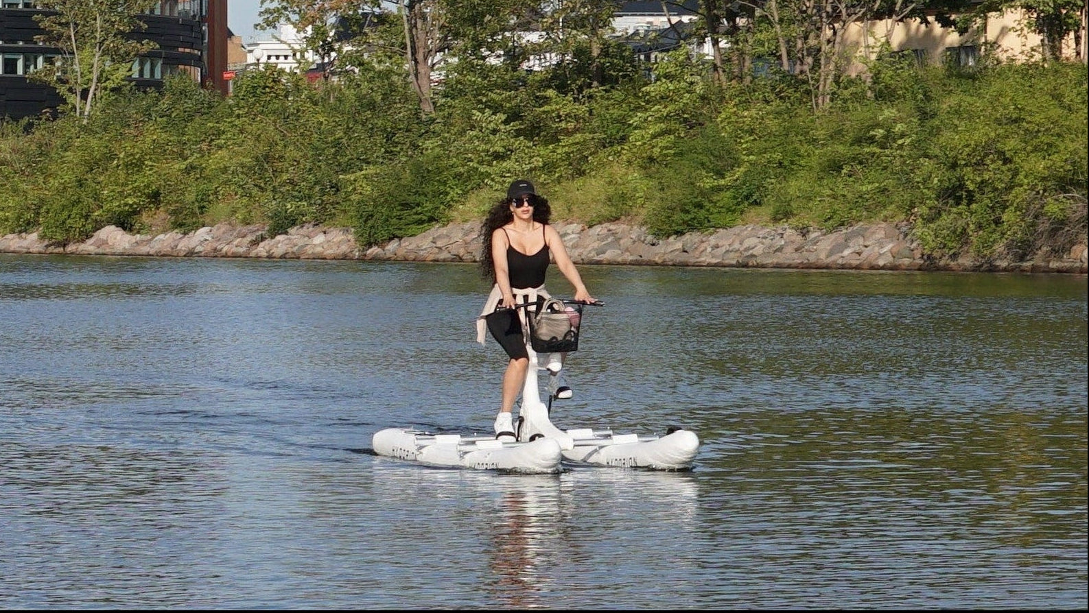 A person stands on a water bike on a calm river with trees, modern buildings, and a blue sky in the background.