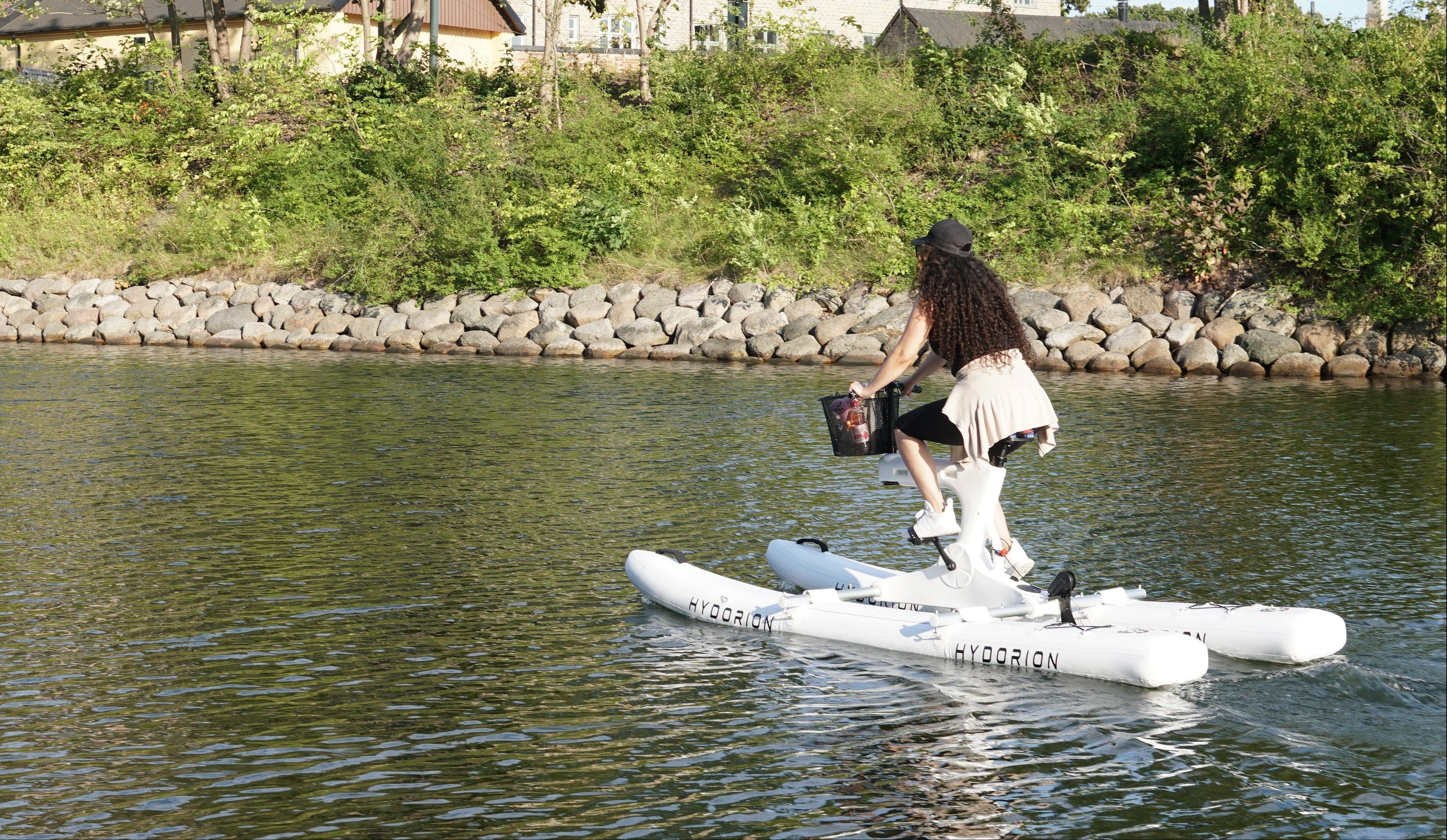 A woman sits on a white water bike on a calm lake, with a stone and grassy shoreline and houses in the background.